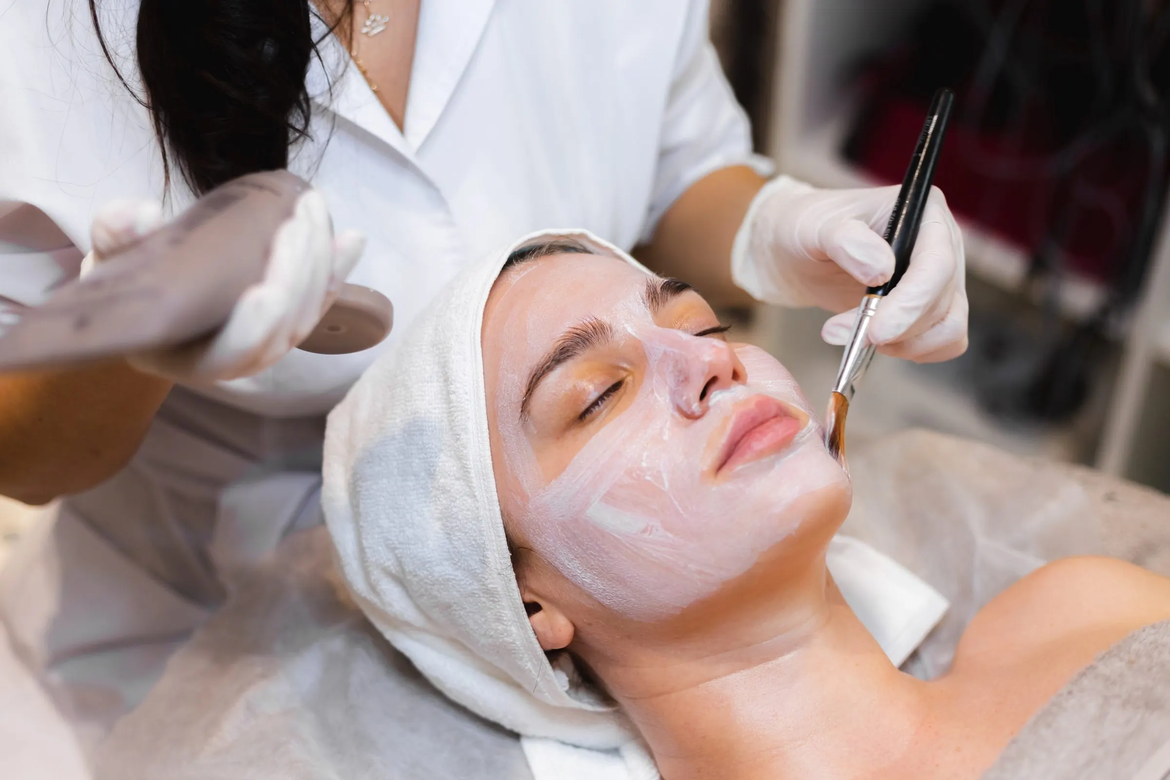 Esteticista con guantes aplicando una mascarilla con brocha a una mujer con turbante de toalla
